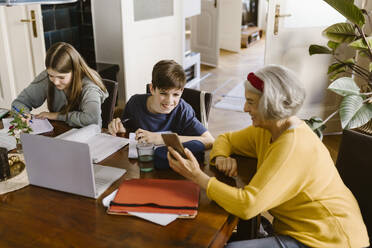 High angle view of senior woman showing smart phone to grandson studying at home - MASF43650