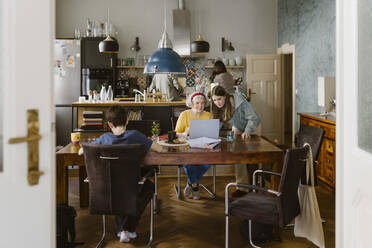 Grandmother using laptop with granddaughter at dining table in home seen through doorway - MASF43656