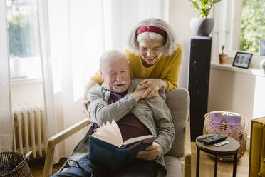 Happy senior woman with arm around man reading book while sitting on chair at home - MASF43668