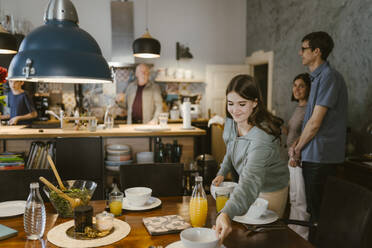 Smiling teenage girl placing bowls on dining table for dinner with family at home - MASF43679