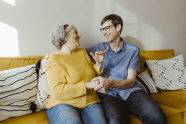 Happy man with arm around senior mother-in-law sitting on sofa at home - MASF43718