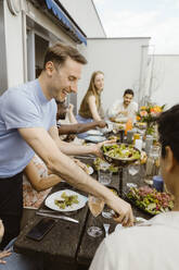 Smiling man serving food to male friend sitting at dining table while celebrating dinner party in balcony - MASF43742