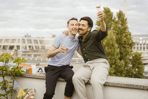 Carefree gay couple toasting drinks while sitting on wall during party in balcony - MASF43761