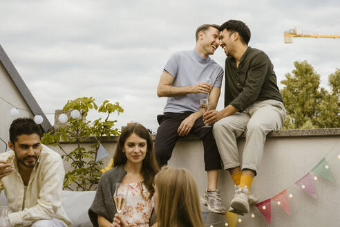 Happy gay couple sitting on wall with friends talking with each other at party in balcony - MASF43764