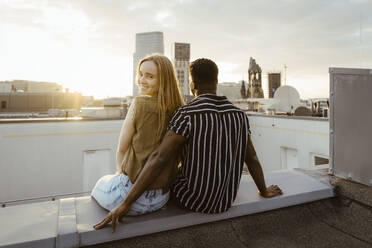 Portrait of smiling woman looking over shoulder while sitting with boyfriend on building terrace - MASF43793