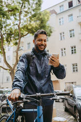 Smiling food delivery person using smart phone while holding bicycle - MASF43811