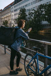 Female delivery person using smart phone while leaning on railing by bicycle on footpath - MASF43851