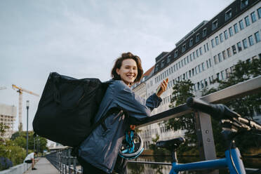 Side view portrait of smiling female delivery person holding smart phone while leaning on railing in city - MASF43852