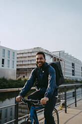 Portrait of smiling young male delivery person cycling on footpath in city - MASF43857