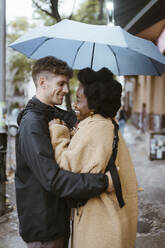 Side view of happy multiracial couple standing under umbrella at street - MASF43979