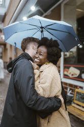 Boyfriend embracing happy multiracial girlfriend under umbrella at street - MASF43980