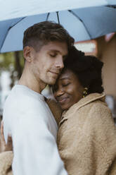 Side view of multiracial romantic couple embracing with eyes closed under umbrella - MASF43981
