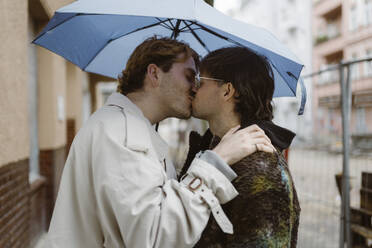 Side view of affectionate gay couple kissing under umbrella at street - MASF43992