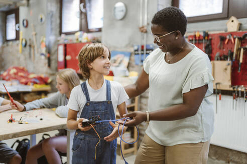 Lächelnde Lehrerin ermutigt Schüler, die ein elektrisches Teil in einem Technik-Workshop in der Schule halten - MASF44149