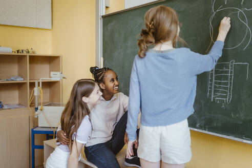 Mädchen haben Spaß mit weiblichen Freund Zeichnung auf Kreidetafel im Klassenzimmer - MASF44177
