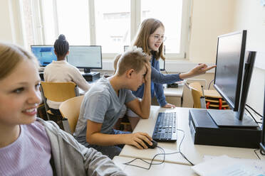Girl pointing on monitor screen with male friend sitting at desk in classroom at school - MASF44192