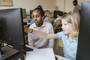 Girl pointing at computer monitor while sitting with female friend in classroom at school - MASF44204