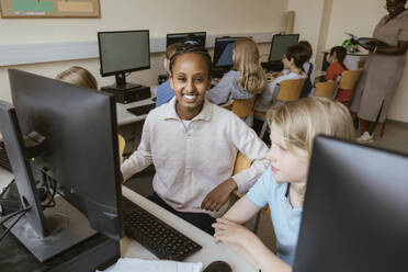 Portrait of smiling girl sitting on chair with friends in computer classroom at school - MASF44205