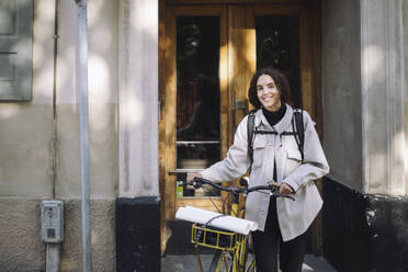 Portrait of smiling female architect standing with bicycle at building entrance - MASF44240