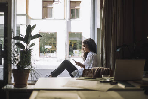 Seitenansicht einer Architektin, die ein Smartphone benutzt, während sie auf der Fensterbank im Büro sitzt - MASF44272