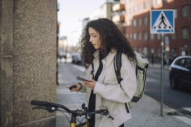 Female construction freelancer pairing bluetooth headphones with smart phone while standing by bicycle near wall at stre - MASF44310