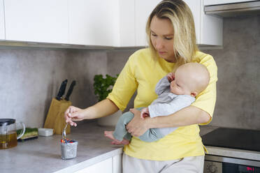 A young mother with European appearance and long blond hair stays at the kitchen, holds a baby in her arms and mixes with a teaspoon of chia yogurt - NJAF00976