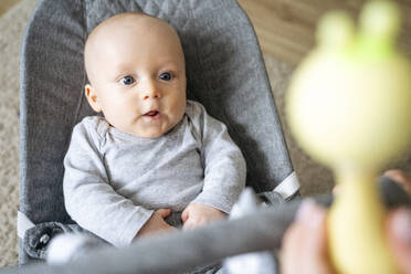 A 5 years old baby is sitting in a grau baby rocking chair at home, someone is showing a toy to him - NJAF00980