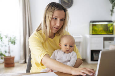 A young mother with European appearance and long blond hair sits at home, holds a baby in her arm and works on a lapton at the table, next to her there are a paper, a pen and a smartphone - NJAF00986
