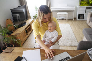 A young mother with European appearance and long blond hair sits at home, holds a baby in her arm and works on a lapton at the table, next to her there are a paper, a pen and a smartphone - NJAF00987