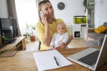 A young mother with European appearance and long blond hair sits at the table, holds a baby in her arm and talks on her smartphone, next to her there are a laptop - NJAF00988