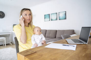 A young mother with European appearance and long blond hair sits at the table, holds a baby in her arm and talks on her smartphone, next to her there are a laptop - NJAF00989