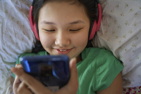 USA. NY, Cute carefree girl using smartphone and headphones while relaxing on a bed - AZF00605