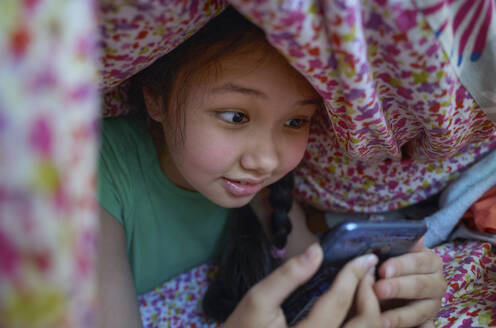 USA. NY, Cute carefree girl using smartphone while relaxing on a bed under the blanket - AZF00606