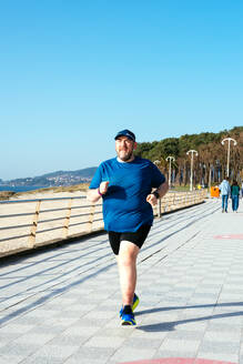 Mann in blauem Hemd und Mütze, der an einem klaren Tag am Meer joggt und für Fitness und Gesundheit wirbt - ADSF54877