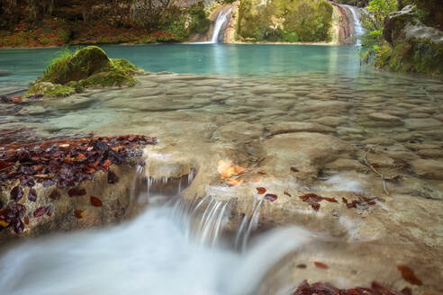 Ruhiges Wasser stürzt in Kaskaden über die mit Herbstblättern geschmückten Felsformationen des Urederra-Flusses vor der Kulisse eines ruhigen Wasserfalls - ADSF55084