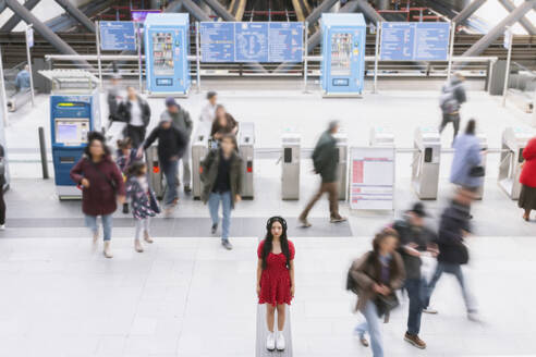 Eine junge Frau in einem roten Kleid bleibt regungslos stehen, während unscharfe Gestalten in einer belebten Bahnhofsumgebung an ihr vorbeirauschen, um das Konzept der Ruhe im Chaos zu veranschaulichen - ADSF55108