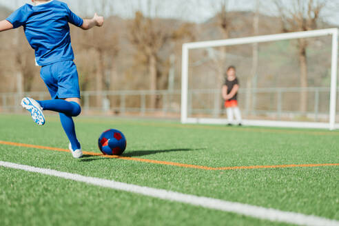 A young soccer player in blue is about to kick a red and blue ball on a vibrant green field, with a goalkeeper in the background - ADSF55144