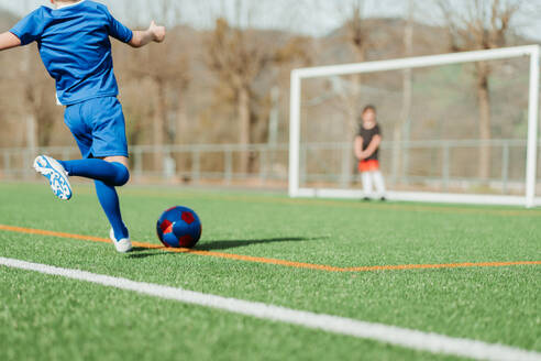 Ein junger Fußballspieler in Blau ist dabei, einen roten und blauen Ball auf einem leuchtend grünen Feld zu schießen, mit einem Torwart im Hintergrund - ADSF55144