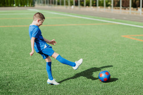 Ein Junge in einem blauen Trikot kickt einen roten Fußball auf einem saftig grünen Kunstrasenplatz und zeigt dabei Entschlossenheit und Sportsgeist - ADSF55147