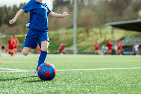 Teilansicht eines jungen Fußballspielers in blauer Uniform, der einen lebendigen Ball auf einem üppigen grünen Spielfeld dribbelt - ADSF55151