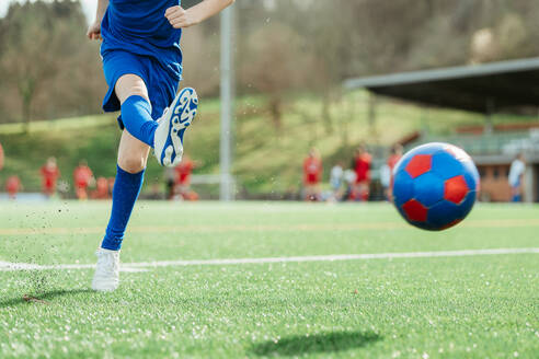 Nahaufnahme eines Fußballspielers in Blau, der einen roten und blauen Fußball auf einem üppigen grünen Feld kickt, mit Spielern im Hintergrund - ADSF55152