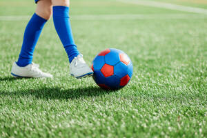Die Beine eines Fußballspielers in blauen Socken und weißen Schuhen, der im Begriff ist, einen roten und blauen Fußball auf einem üppigen grünen Feld zu treten - ADSF55153