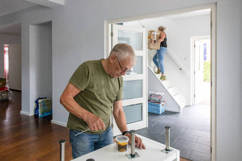 Älterer Mann mit Brille konzentriert sich auf eine Aufgabe in einem modernen Haus, während jemand die Treppe im Hintergrund hinaufsteigt. - ISF26627