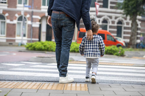 Ein Elternteil und ein Kind halten sich an den Händen, während sie an einem bewölkten Tag die Straße überqueren. - ISF26739