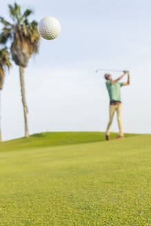 Ein Golfspieler mit einzigartigen Dreadlocks wird an einem sonnigen Tag mitten im Schwung aufgenommen, wie er den Ball hoch in Richtung der Palmen schlägt. - ADSF55183