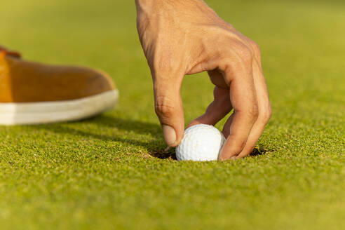 Abgeschnittene, nicht erkennbare Hand eines Golfspielers mit Dreadlocks, der vorsichtig einen weißen Golfball auf dem grünen Rasen platziert, was einen Moment der Präzision im Spiel illustriert. - ADSF55186