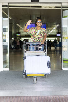 Ein fröhlicher Reisender winkt zur Begrüßung und schiebt einen Gepäckwagen am Flughafen, um die Aufregung des Reisens und der Ankunft festzuhalten - ADSF55196