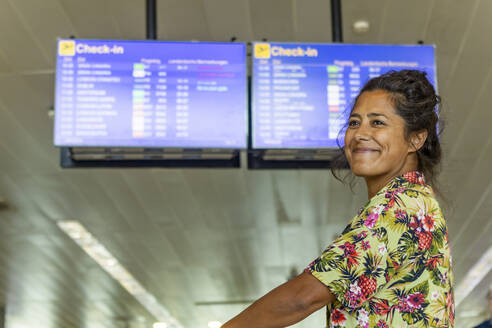 Eine fröhliche Frau steht an einem Flughafen-Check-in-Bereich, mit Fluginformationsanzeigen im Hintergrund - ADSF55198