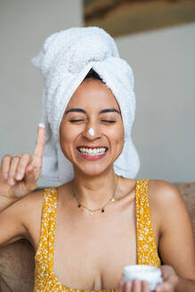 Eine fröhliche Frau mit einem Handtuch auf dem Kopf trägt Gesichtscreme auf und genießt ihre morgendliche Schönheitspflege - ADSF55221