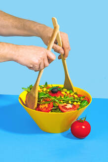 Hands toss a vibrant fresh salad using wooden spoons in a yellow bowl on a blue background - ADSF55245